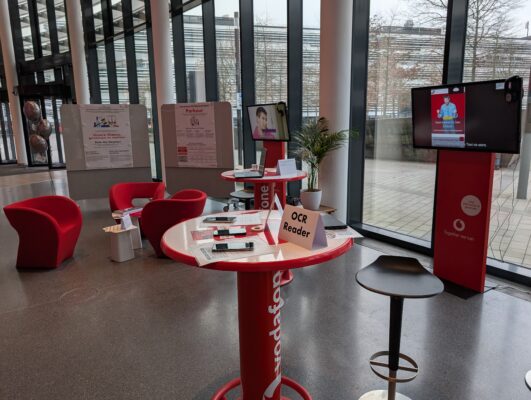 Bild der Lounge, Aufnahme der Parkour-Ecke. Diese besteht aus 2 Aufstellern, 2 Standtischen, 2 TV-Geräten und einer Sitzecke mit Sesseln für 3 Personen. Es liegen einige Smartphones, Tablets und Laptops aus. Es sind keine Personen im Bild zu sehen.