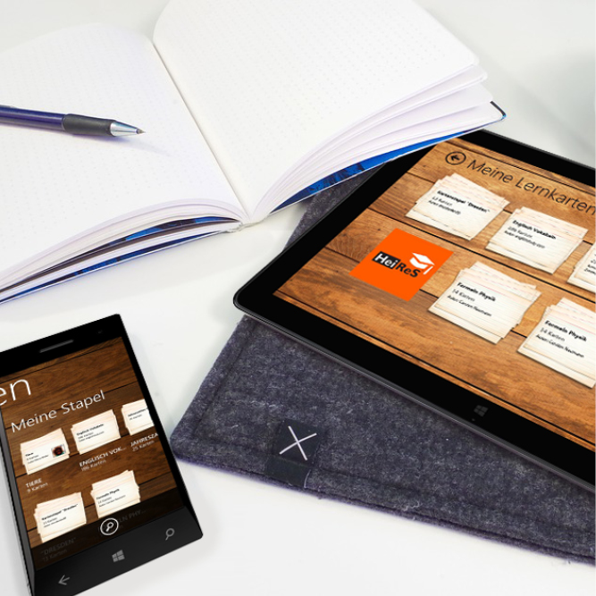 Nahaufnahme eines Tisches, auf dem ein geöffnetes Notizbuch samt Stift, ein Smartphone und ein Tablet liegen, Smartphone und Tablet zeigen das Lernkartensystem.