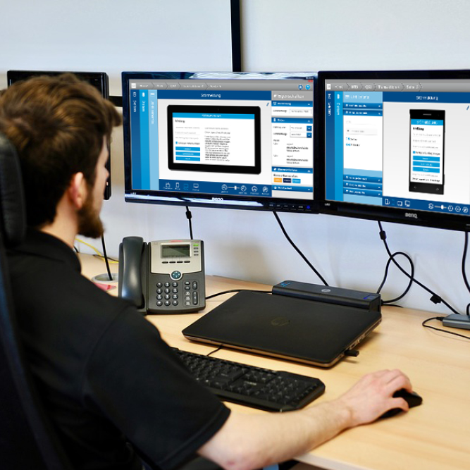 Nahaufnahme Arbeitsplatz mit Telefon, Laptop, Tastatur und mehreren Monitoren, die Monitore zeigen die neue Oberfläche.